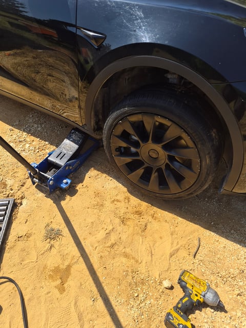 Close-up of a Tesla Model Y black Gemini alloy wheel and Michelin tire with the car held on a blue floor jack at the pinch-weld lift point, cordless impact wrench and socket rail on the ground during an on-site EV tire service by All Hours Roadside in DFW