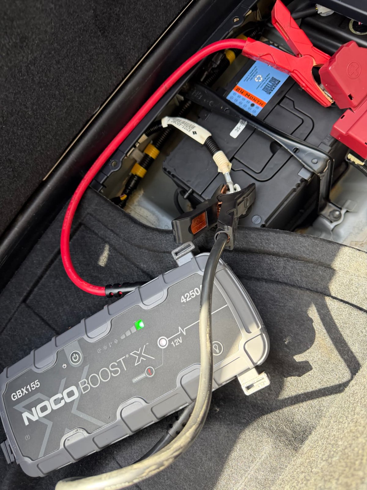 NOCO Boost X GBX155 jump pack clamped to a trunk-mounted 12V car battery with red positive cable and ground strap during a mobile jump start in Addison TX by All Hours Roadside