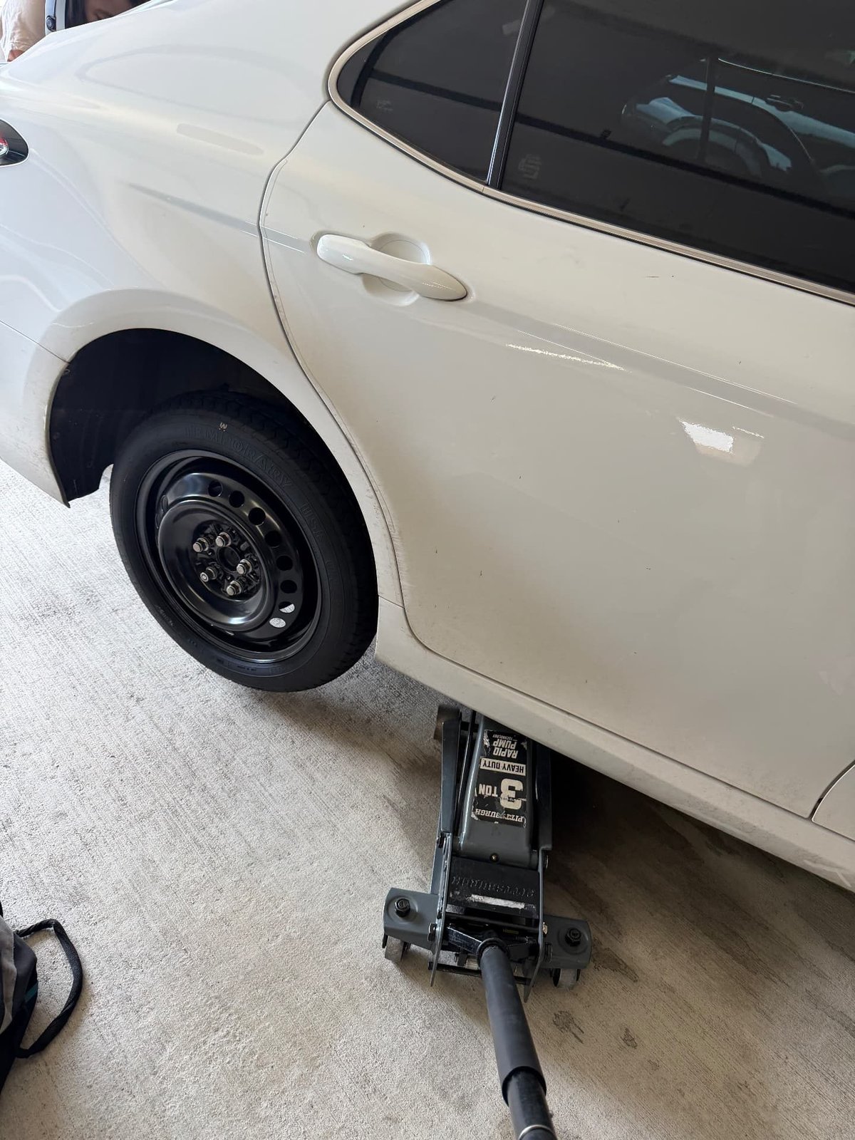 White sedan raised on a 3-ton Pittsburgh floor jack with the factory temporary spare wheel mounted on the rear hub during a mobile tire change in Irving TX by All Hours Roadside