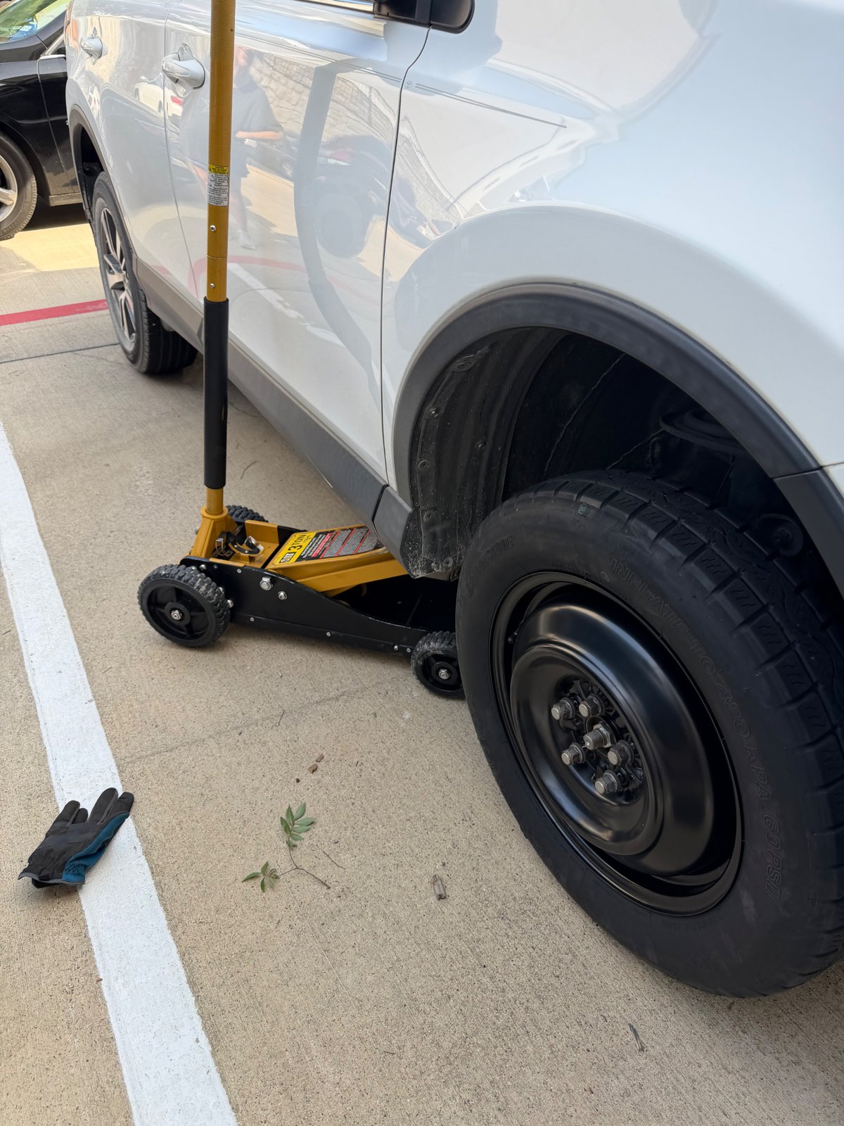 All Hours Roadside spare tire change on a white Toyota RAV4 in a Euless, TX parking lot — 3-ton yellow Pittsburgh floor jack supporting the vehicle, black steel spare wheel installed on the rear passenger side.