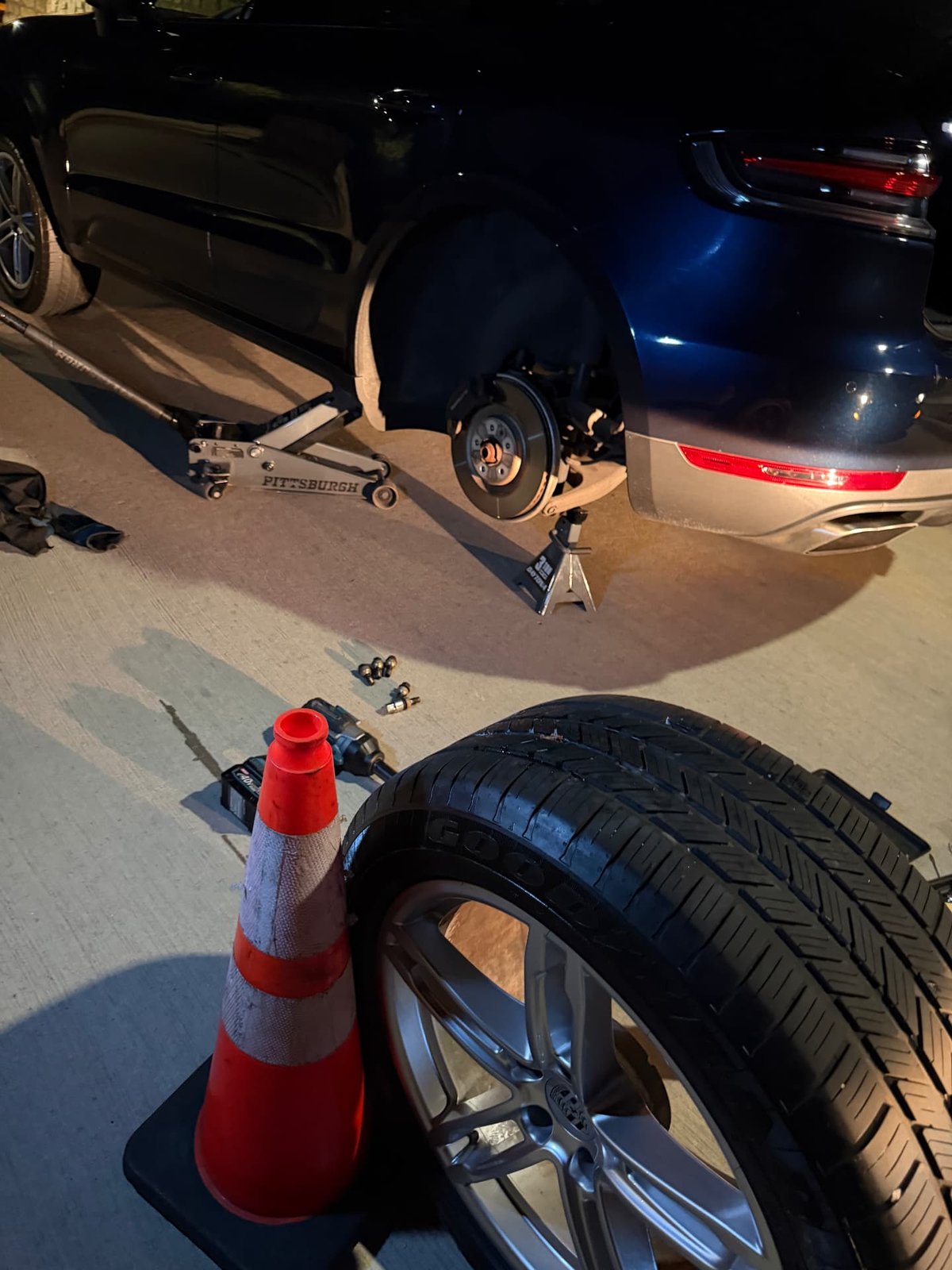 Dark Porsche SUV raised on a floor jack and jack stand at night with the rear wheel removed, cordless impact gun, lug bolts and a Goodyear tire on an alloy wheel staged beside a safety cone during a mobile tire change in Addison TX by All Hours Roadside