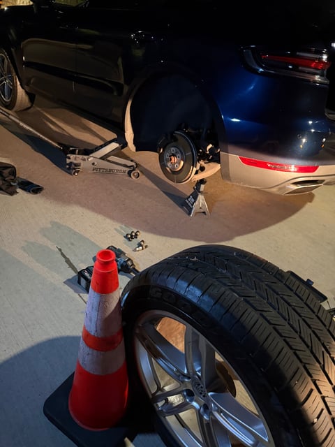 Dark Porsche SUV raised on a floor jack and jack stand at night with the rear wheel removed, cordless impact gun, lug bolts and a Goodyear tire on an alloy wheel staged beside a safety cone during a mobile tire change in Addison TX by All Hours Roadside