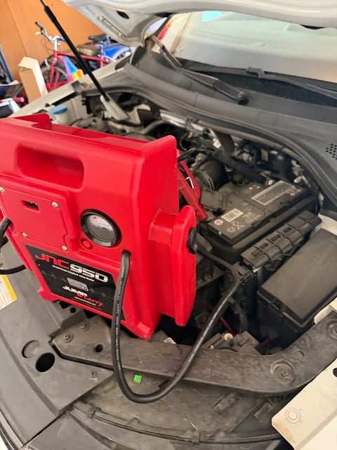 JNC950 lithium jump pack clamped to a dead battery on a white sedan inside an Uptown Dallas TX parking garage — jump start by All Hours Roadside