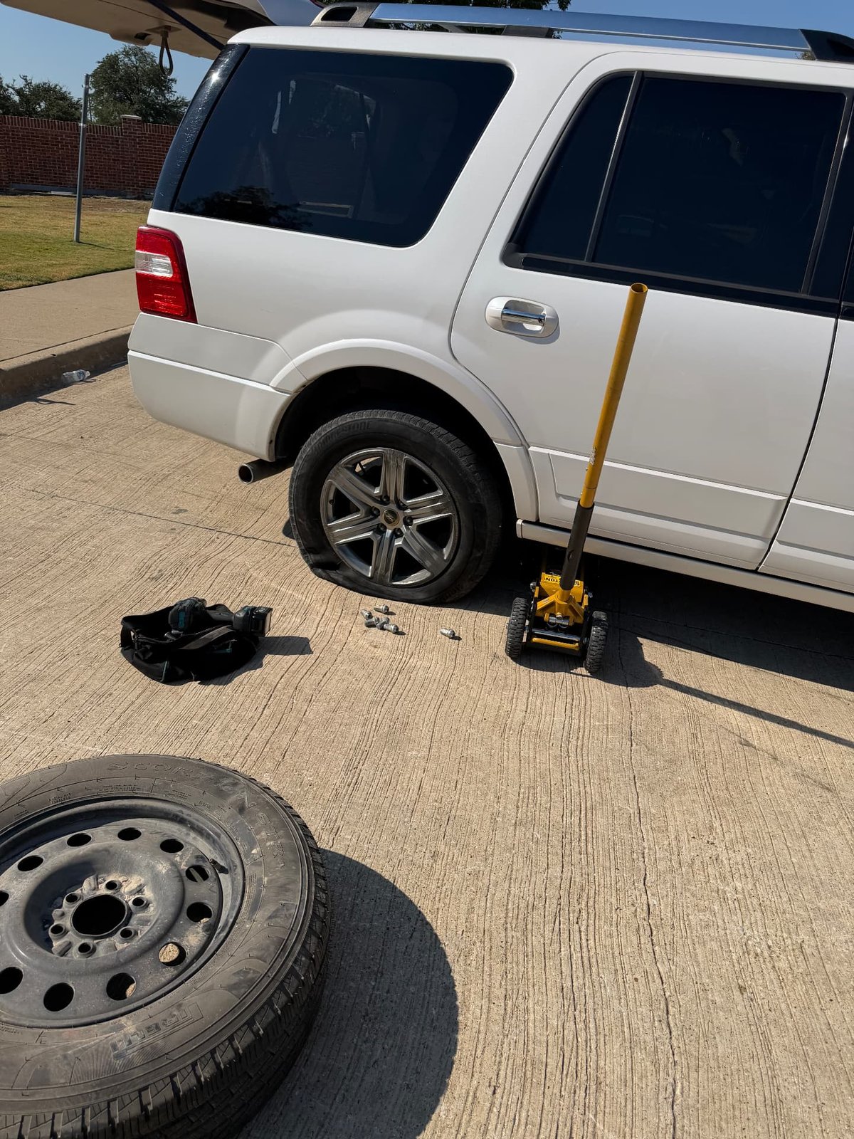 White Ford Expedition raised on a yellow low-profile floor jack in a Fort Worth TX driveway with the rear wheel removed and a full-size steel spare on the ground during a mobile tire change by All Hours Roadside