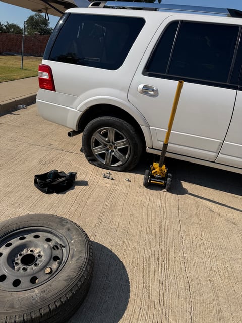 White Ford Expedition raised on a yellow low-profile floor jack in a Fort Worth TX driveway with the rear wheel removed and a full-size steel spare on the ground during a mobile tire change by All Hours Roadside