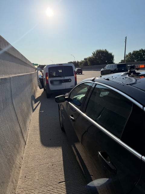 White Ram ProMaster City cargo van stopped on a DFW highway shoulder against a concrete barrier wall with its rear door open, and an amber light-bar response vehicle parked behind it blocking the lane during an emergency mobile tire change by All Hours Roadside
