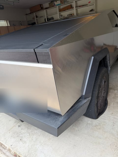 Silver Tesla Cybertruck in a garage with a visibly deflated rear tire before mobile tire service; license plate obscured