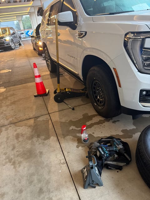 White GMC Yukon raised with a floor jack and supported by a jack stand beside a safety cone during a mobile tire change by All Hours Roadside
