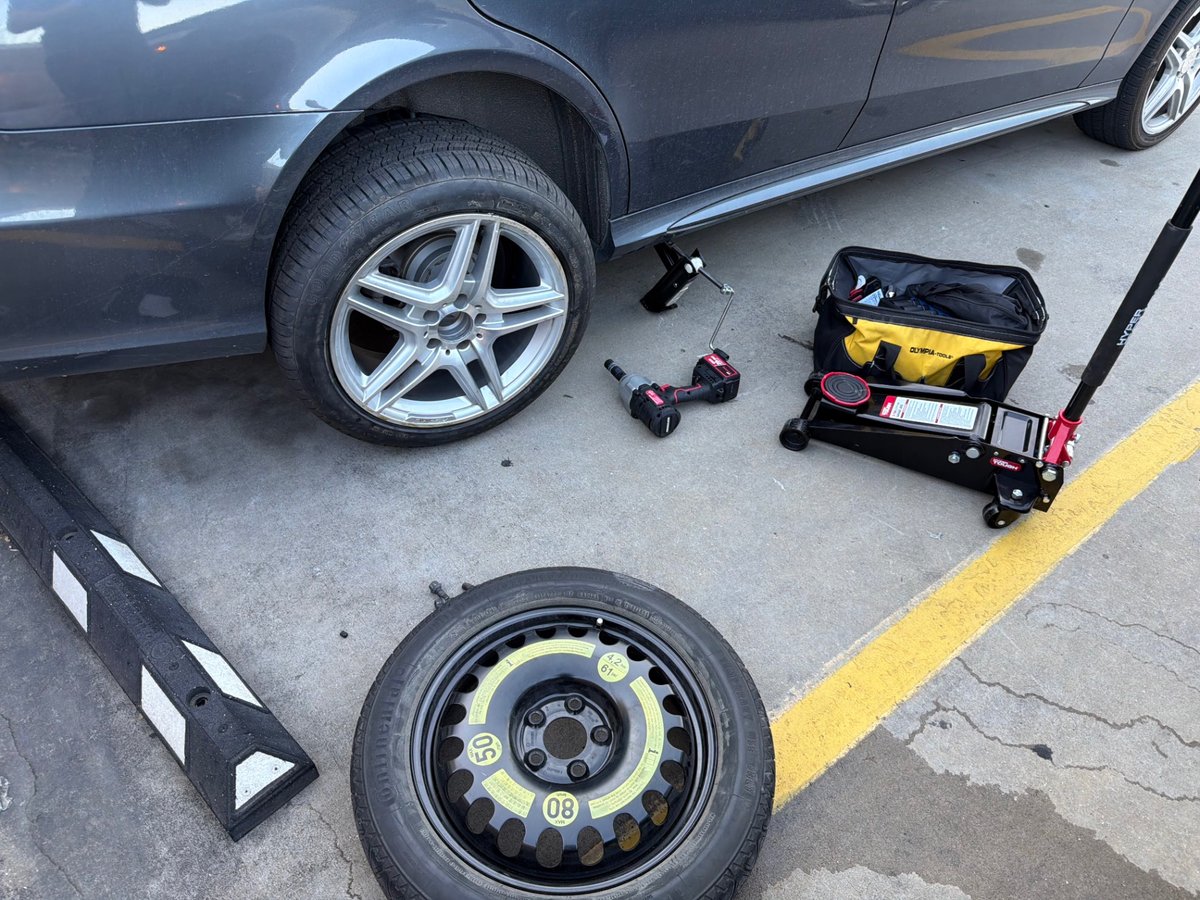 Gray Mercedes-Benz with AMG-style alloy wheel removed and set on a parking block, OEM donut spare staged on the ground, Hyper Tough 3-ton floor jack and cordless impact in a clean Austin TX parking lot — All Hours Roadside mobile tire change