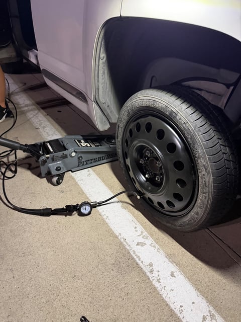 Black steel donut spare tire mounted on a silver sedan lifted on a Pittsburgh floor jack at night in a Flower Mound TX parking lot, with an inflator and pressure gauge reading the spare — All Hours Roadside mobile tire change