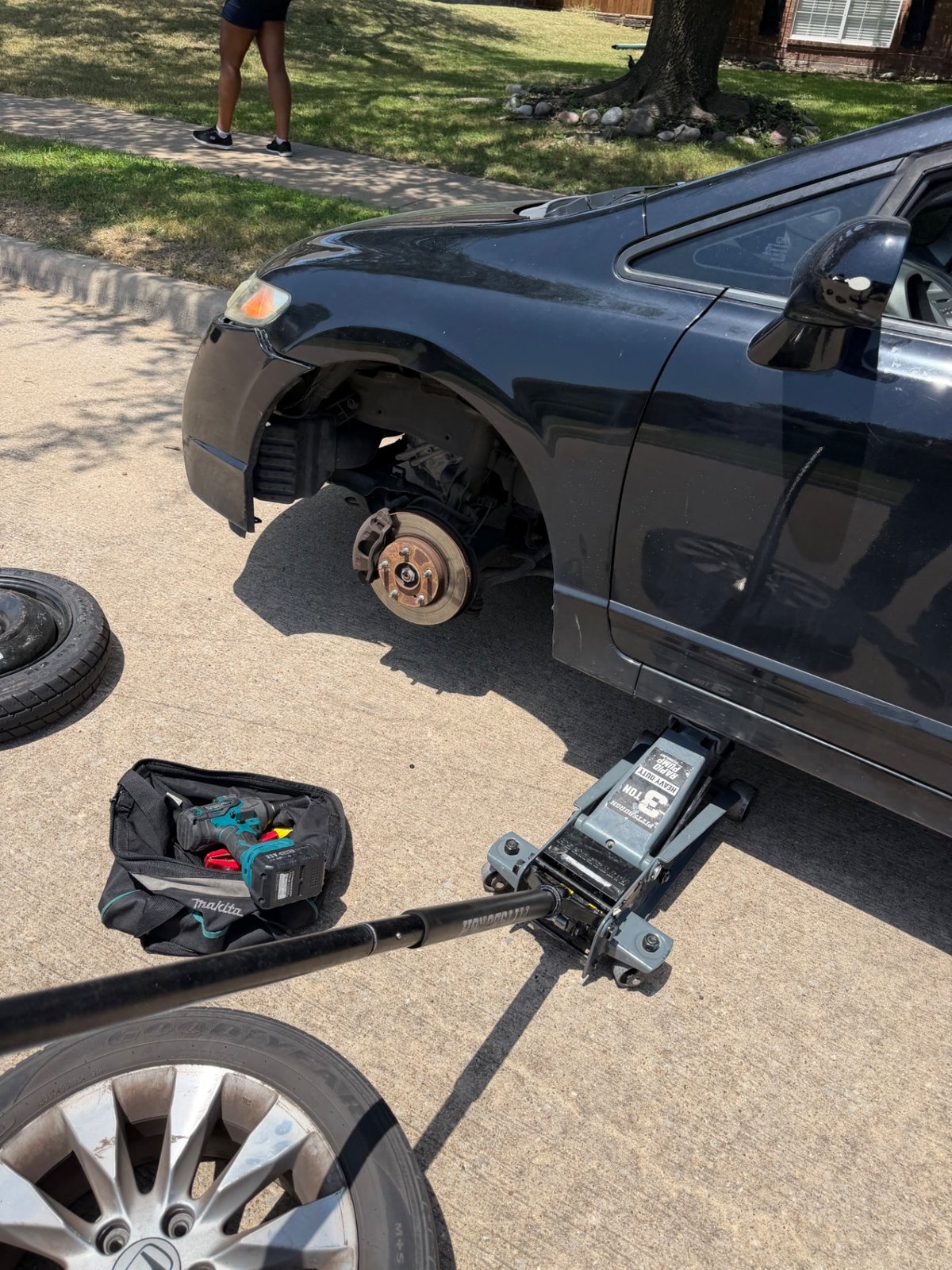 Black Honda Civic lifted on a Pittsburgh 3-ton heavy-duty rapid pump floor jack in a Carrollton TX driveway with the front driver-side wheel removed, Makita tool bag with cordless impact staged curbside, spare and alloy wheel on the ground — mobile tire change by All Hours Roadside