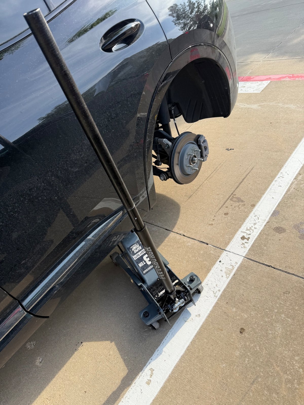 Black SUV raised on a Pittsburgh 3-ton rapid-pump floor jack with rear wheel removed during a roadside spare tire change in a Grand Prairie TX parking lot — All Hours Roadside mobile tire service