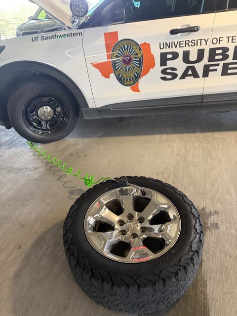 Full-size Ram truck wheel with a chrome alloy rim and all-terrain tire laid on the concrete of a Dallas campus parking deck, green coiled air hose and inflator chuck attached to the valve stem, with a white university public safety Ford Explorer patrol SUV on a black steel spare wheel in the background during a mobile fleet tire change by All Hours Roadside