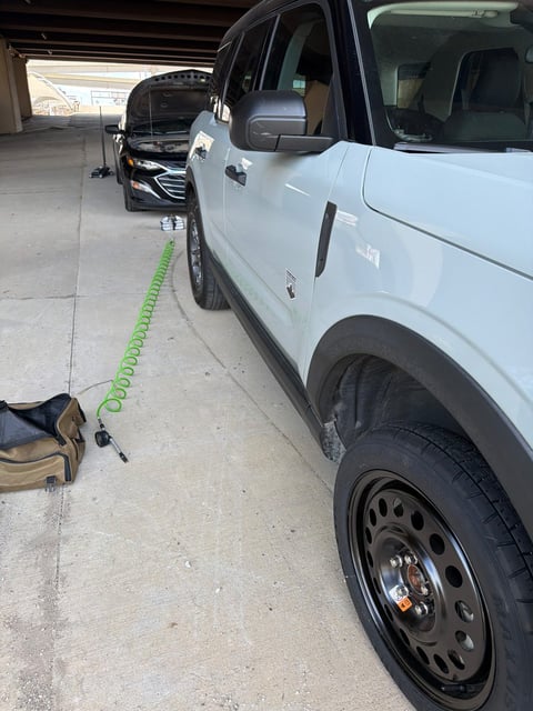 White Ford Bronco Sport Big Bend parked inside a covered DFW Airport terminal parking garage with the front passenger wheel replaced by a black steel donut spare, green coiled air hose and inflator staged on the concrete during a mobile tire change by All Hours Roadside at DFW Airport