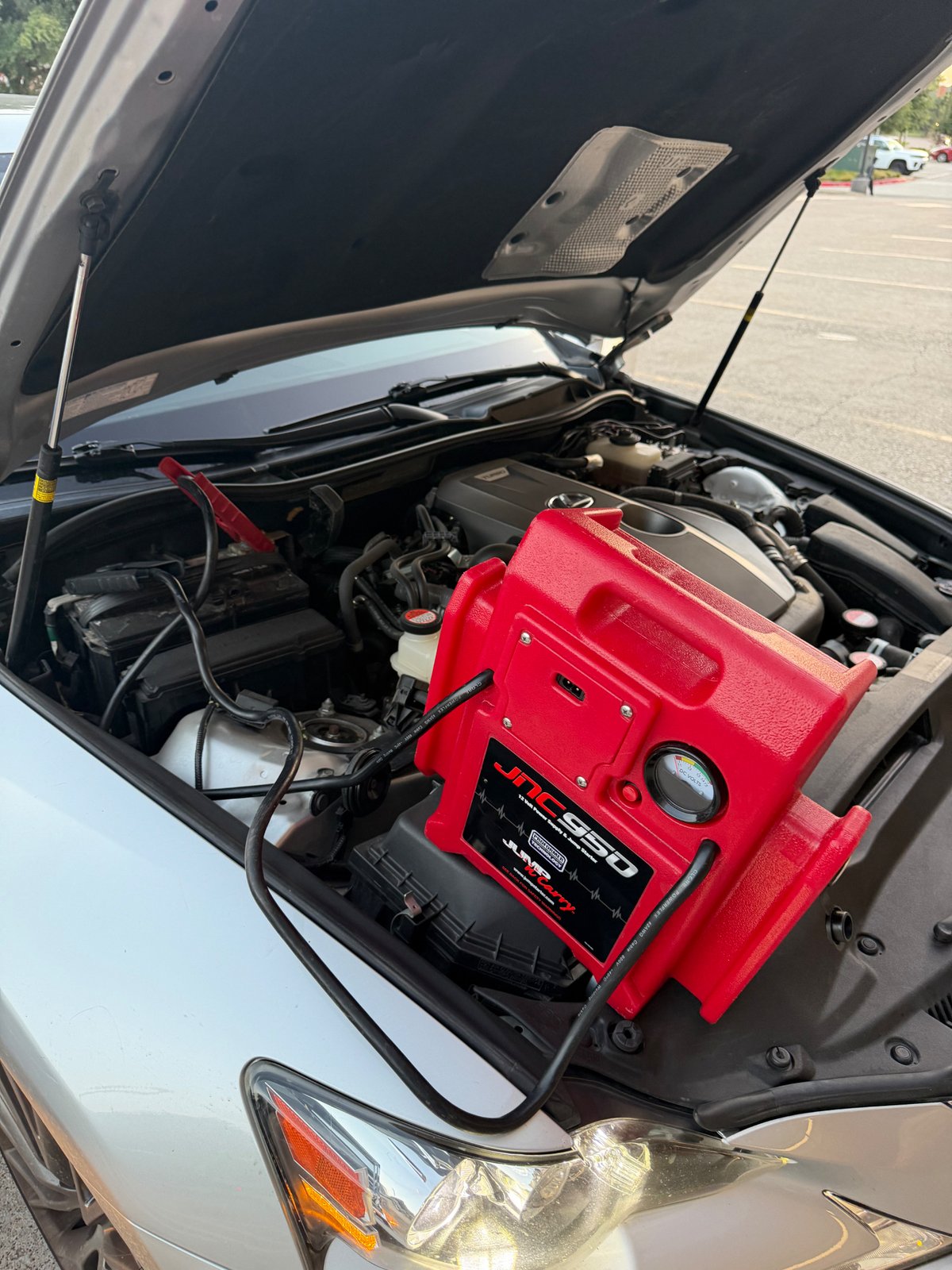 Red Clore JNC950 Jump-N-Carry commercial jump pack clamped to the 12V battery of a silver Toyota Camry with the hood up in a Fort Worth TX parking lot — All Hours Roadside mobile jump start