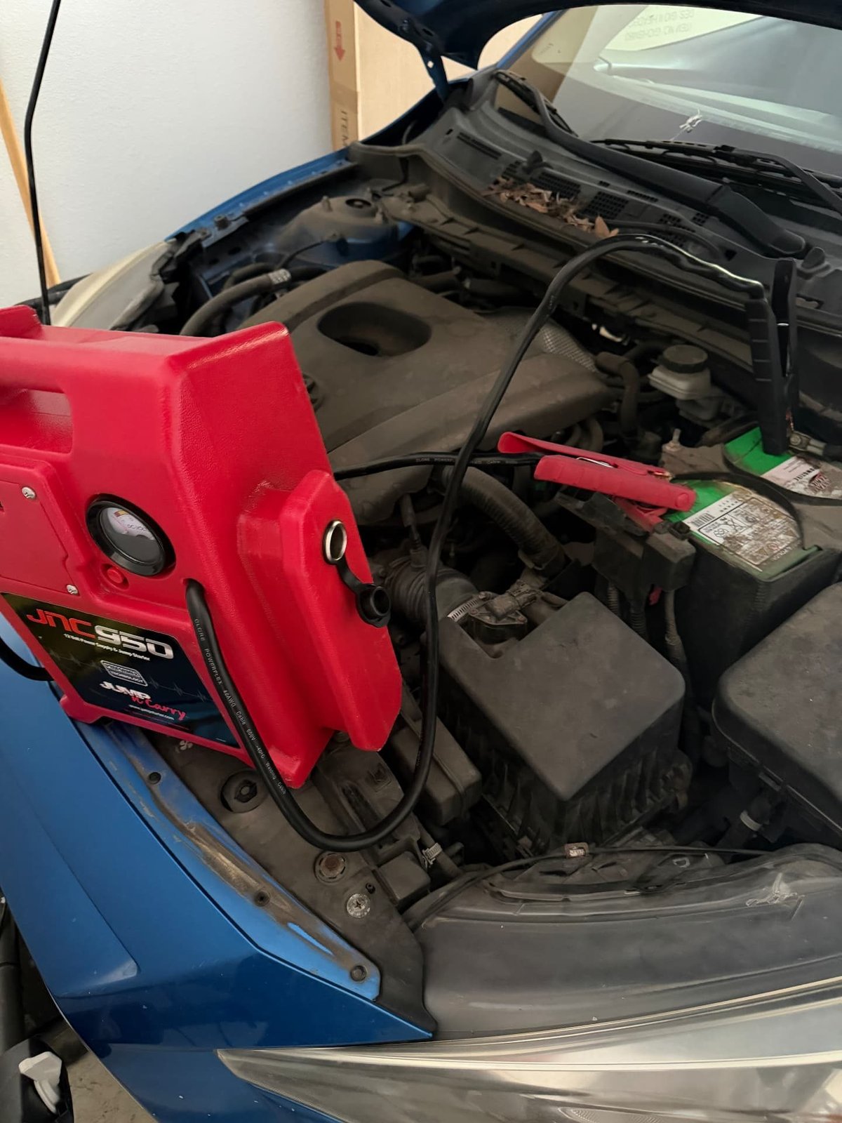 Red Clore JNC950 commercial jump pack with the red clamp on the positive post of a green-top OEM battery in a blue sedan during a mobile jump start in Mansfield TX by All Hours Roadside