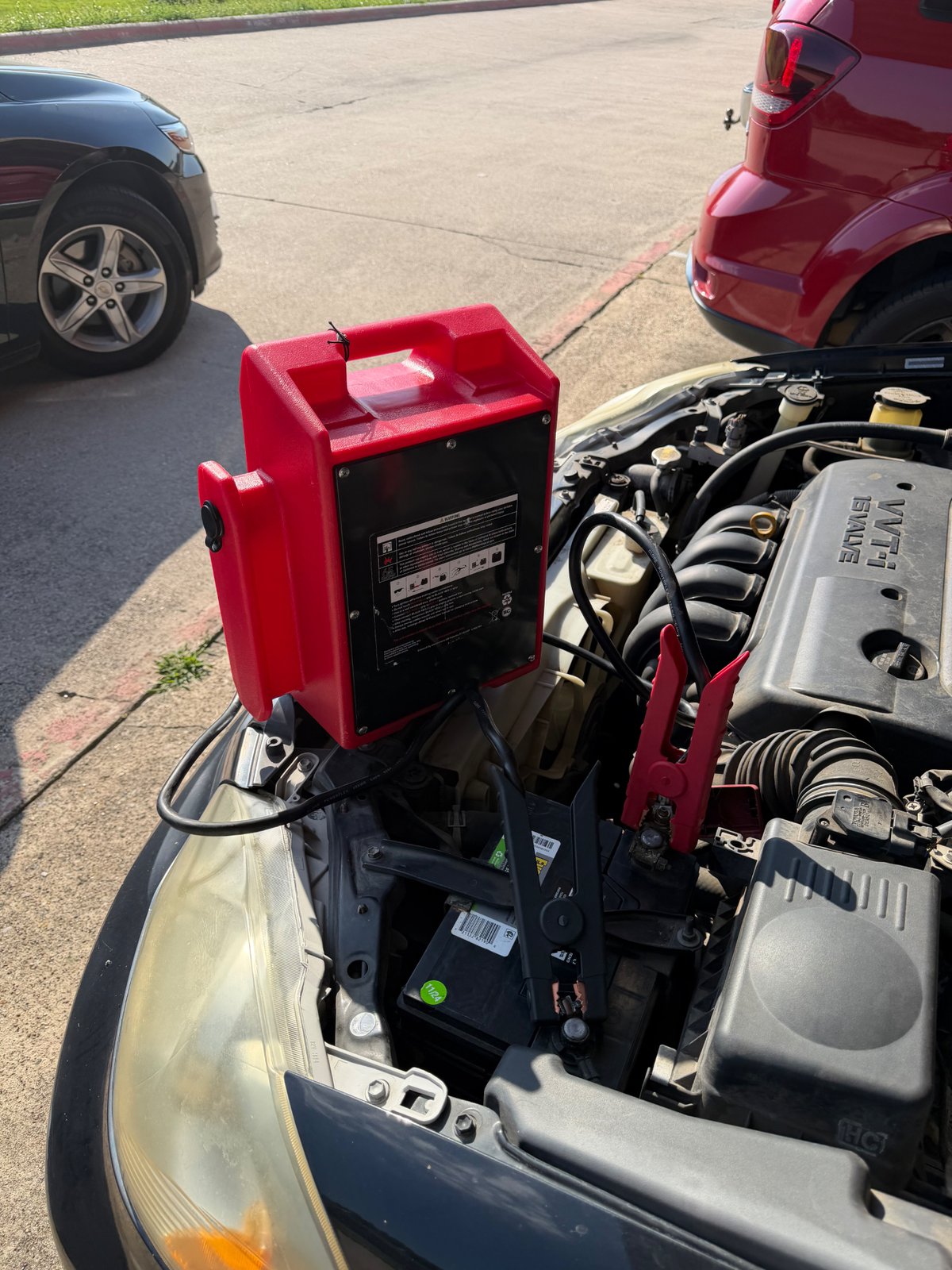 Top-down view of a red commercial jump pack balanced on the core support of a Toyota Corolla in Mesquite TX with red clamps on the OEM battery terminals near the Town East Mall / I-635 corridor — All Hours Roadside mobile jump start