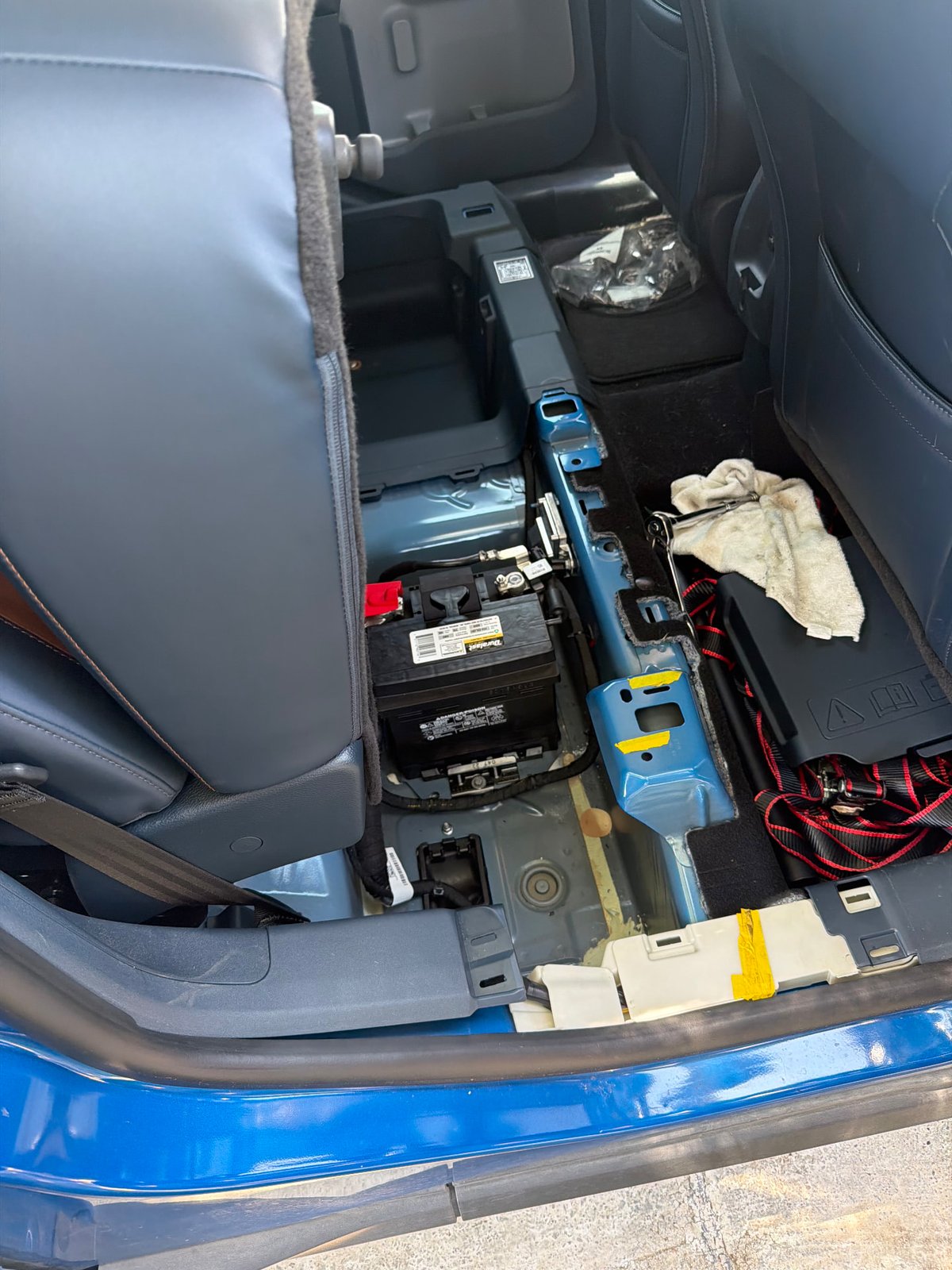 Rear seat cushion removed from a blue 2023 Ford Maverick Hybrid exposing the factory under-seat 12V battery tray during a mobile battery replacement by All Hours Roadside in DFW
