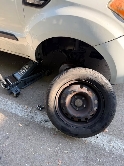 Compact SUV lifted on a 3-ton Pittsburgh floor jack in Plano, TX with a 195/65R15 steel spare tire staged on the pavement and lug nuts set aside for reinstallation by an All Hours Roadside technician