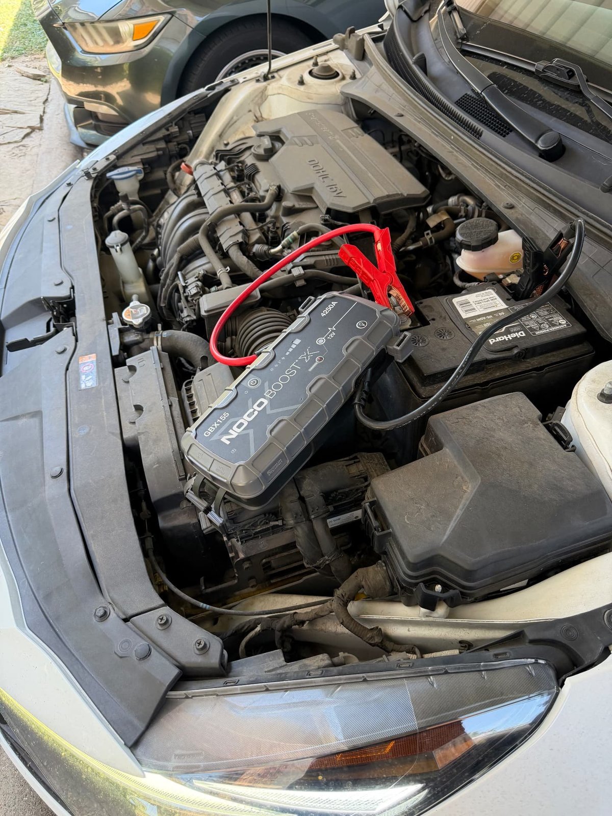 NOCO Boost X GBX155 jump pack clamped to a DieHard AGM battery in the engine bay of a silver car with a DOHC 16V engine cover during a mobile jump start in Keller TX by All Hours Roadside
