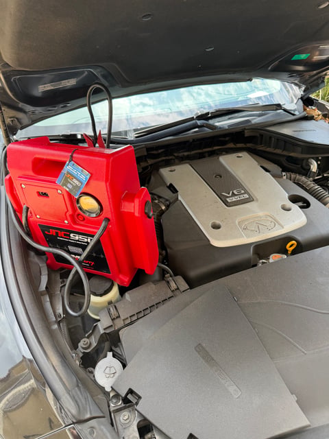 JNC950 professional 12V jump-pack clamped to the OEM battery of an Infiniti V6 VVEL engine during a roadside jump start in a Fort Worth driveway