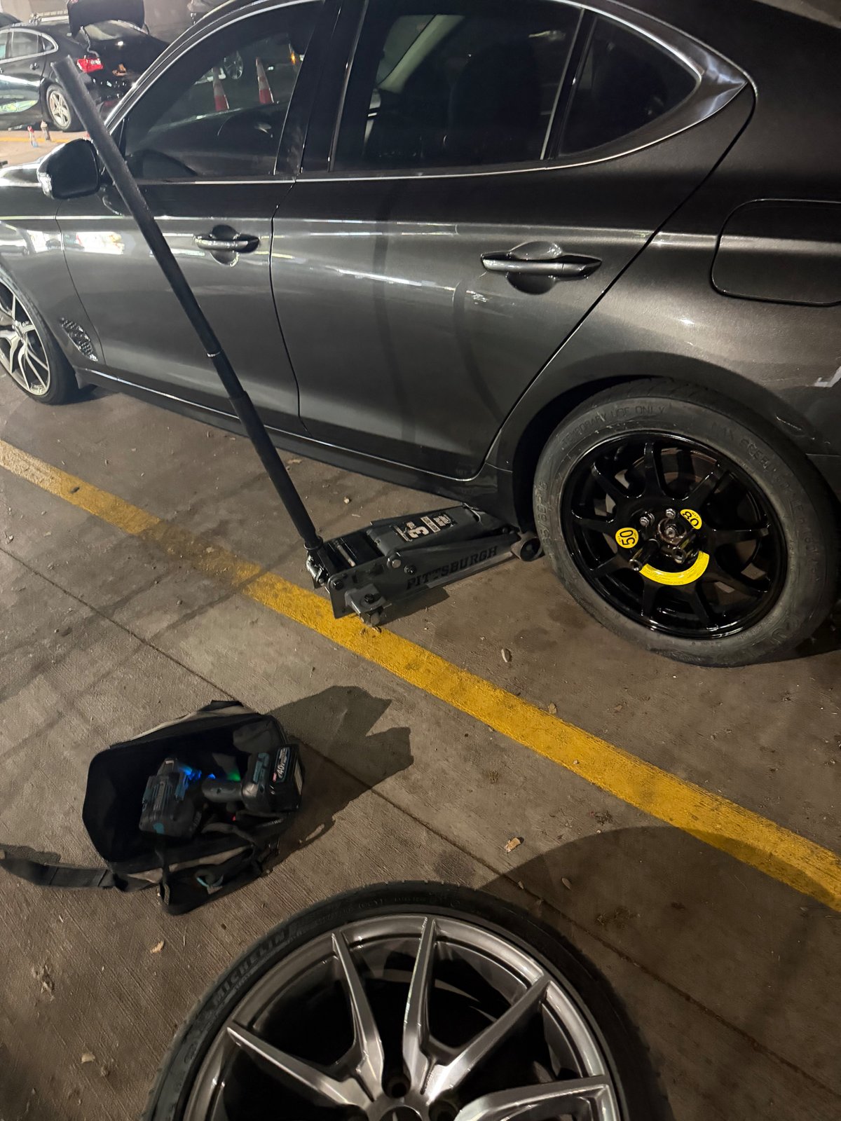 Genesis G70 lifted on a Pittsburgh 3-ton floor jack with a black donut spare mounted and yellow torque-stripe caps on the lug nuts during a mobile spare-tire change in a Frisco TX parking garage — All Hours Roadside