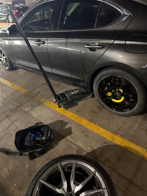 Genesis G70 lifted on a Pittsburgh 3-ton floor jack with a black donut spare mounted and yellow torque-stripe caps on the lug nuts during a mobile spare-tire change in a Frisco TX parking garage — All Hours Roadside