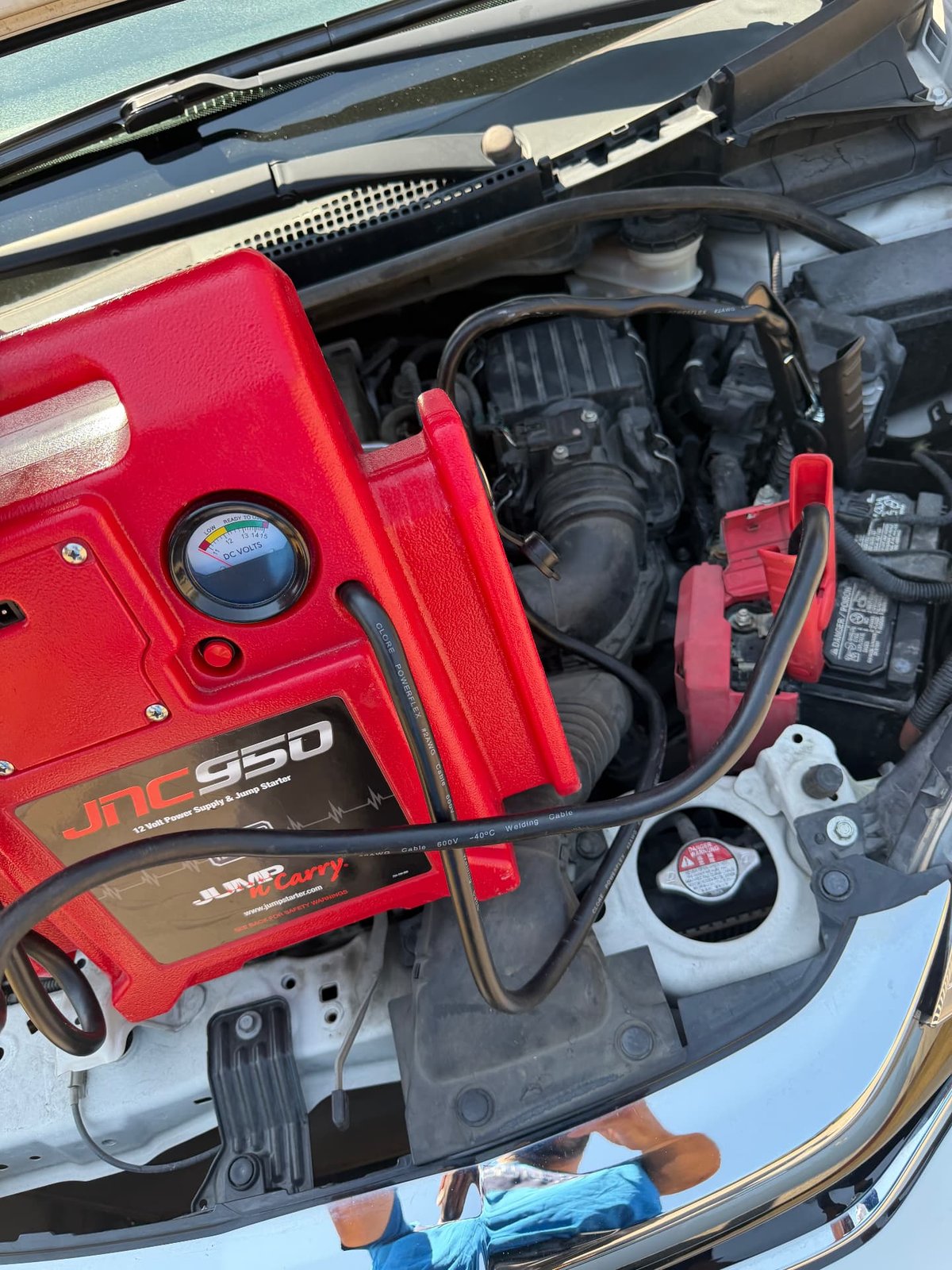 Red JNC950 Jump-N-Carry jump pack clamped to the battery of a white sedan with the hood open during a 24/7 mobile jump start in Fort Worth TX by All Hours Roadside