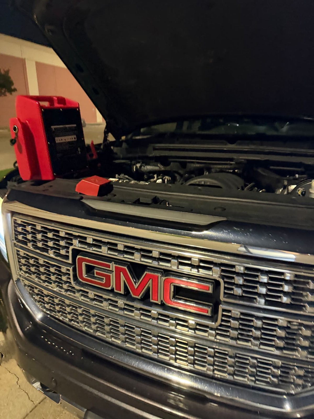 Red heavy-duty portable jump pack set on the radiator support of a black GMC Sierra Denali HD with the hood raised at night in Irving TX — All Hours Roadside mobile jump start