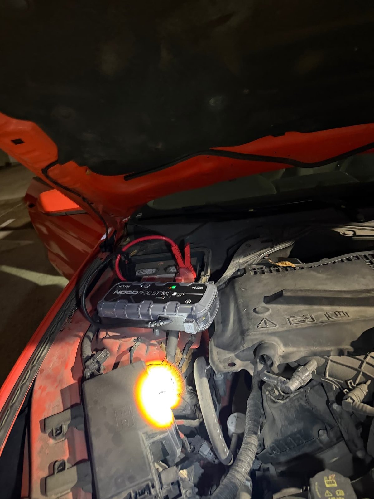 NOCO Boost X jump pack connected to the battery under the open hood of a red car at night, with a work light illuminating the engine bay