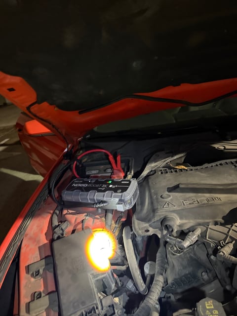 NOCO Boost X jump pack connected to the battery under the open hood of a red car at night, with a work light illuminating the engine bay