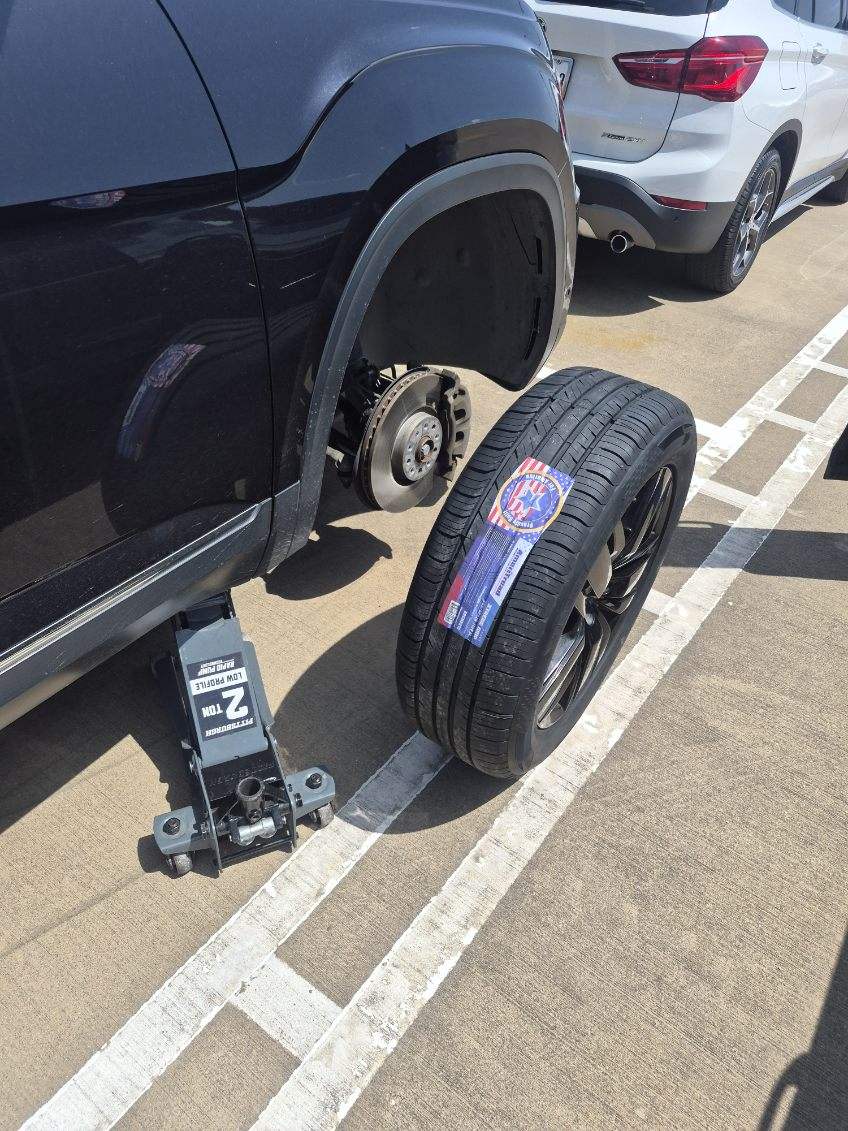 Black Volkswagen Atlas SUV on a Pittsburgh 2-ton floor jack with the rear wheel removed showing the exposed brake rotor and hub, new replacement tire staged next to the vehicle during a mobile tire replacement by All Hours Roadside in Dallas, TX
