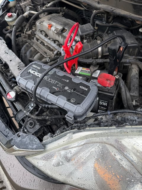 NOCO Boost X GBX155 jump pack with red and black clamps connected to a car battery in a rain-wet engine bay