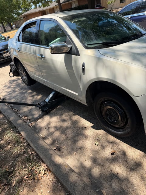 White Lincoln MKZ raised on a black floor jack on a residential street in Irving TX with the temporary donut spare mounted on the rear hub during a mobile tire change by All Hours Roadside