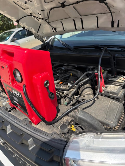 Red JNC950 commercial jump pack set on the radiator support of a white pickup truck with the hood raised, red clamp on the positive battery post, during a mobile jump start in Richardson TX by All Hours Roadside