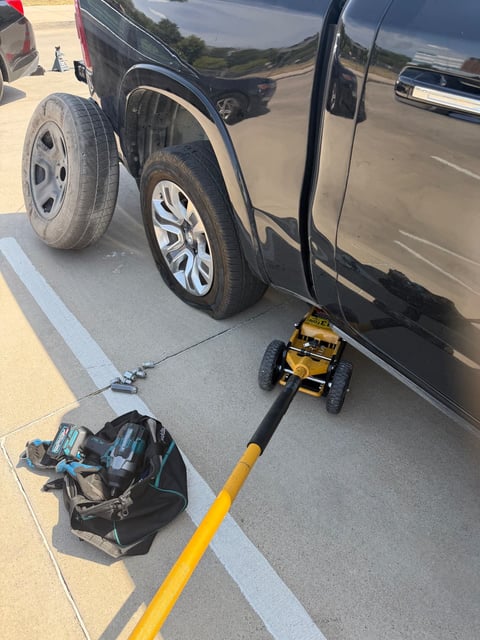 Black Ram 1500 pickup raised on a yellow 3-ton floor jack with the alloy wheel removed and the full-size spare on the pavement during a mobile tire change in Garland TX by All Hours Roadside