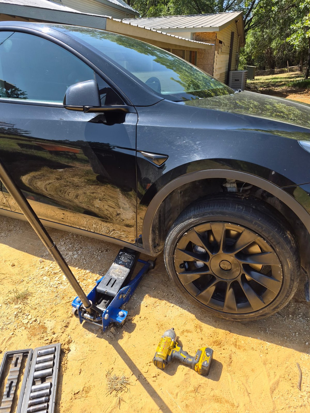 Black Tesla Model Y lifted at the front-right pinch-weld jack point with a blue low-profile floor jack on a gravel driveway in DFW