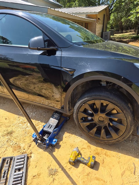 Black Tesla Model Y raised on a blue low-profile floor jack positioned at the factory pinch-weld jack point behind the front wheel, with a cordless impact wrench and socket set staged on the ground during a mobile EV tire change by All Hours Roadside in DFW