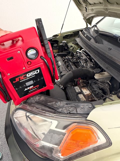 Technician holding a red Clore JNC950 Jump-N-Carry commercial jump pack at the open hood of a tan sedan with the red clamp on the positive battery terminal during a mobile jump start in DeSoto TX by All Hours Roadside