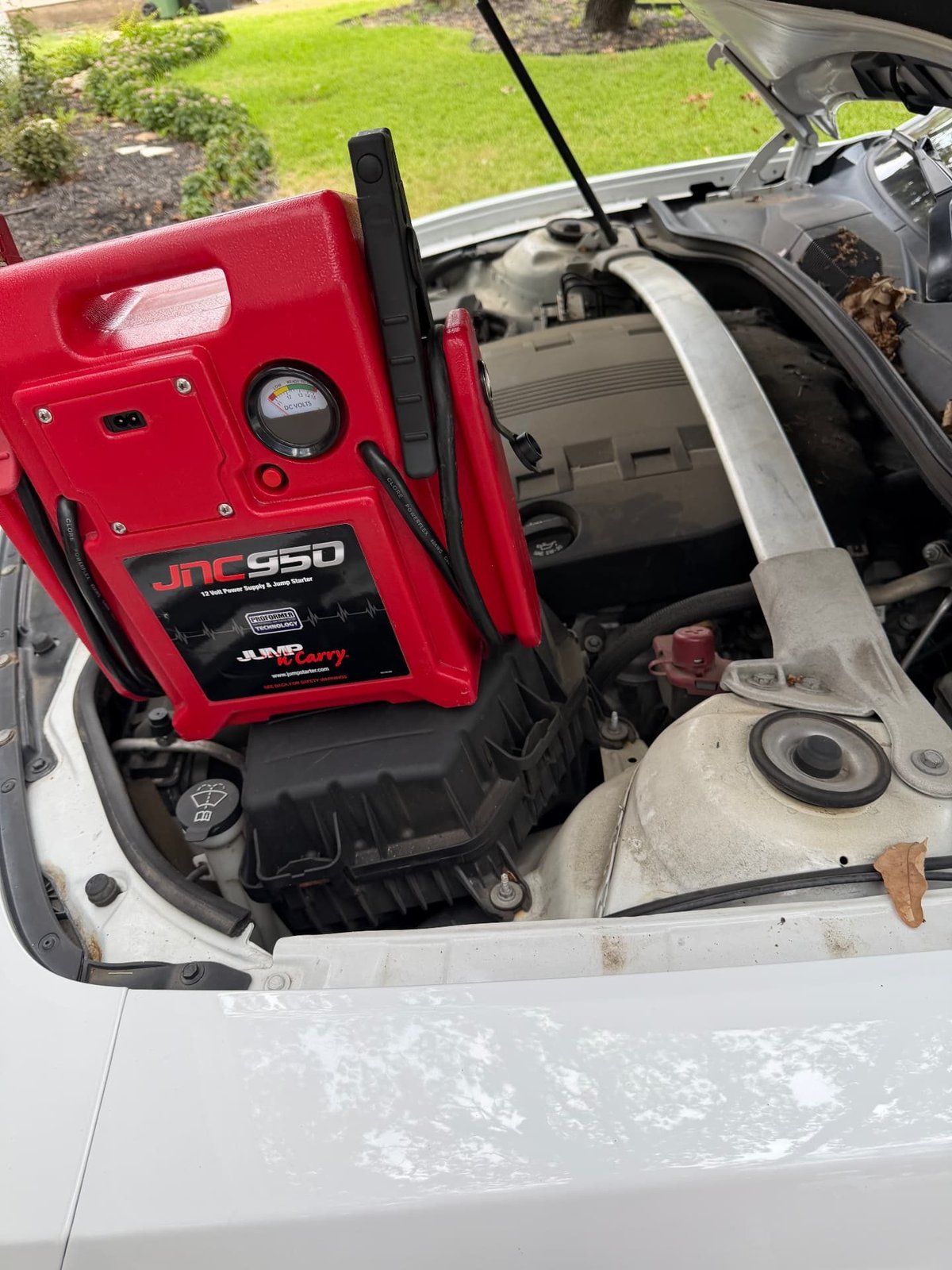 Red Clore JNC950 Jump-N-Carry commercial jump pack propped in the engine bay of a white sedan with the hood raised in a Dallas TX driveway during a 24/7 mobile jump start by All Hours Roadside