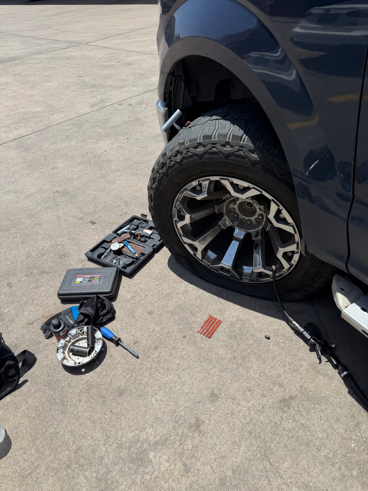 Lifted dark-blue Ford F-150 on chrome wheels with a tire plug kit and portable 12V air compressor re-inflating the front tire on a clean concrete pad in Southlake TX — All Hours Roadside on-vehicle tire repair