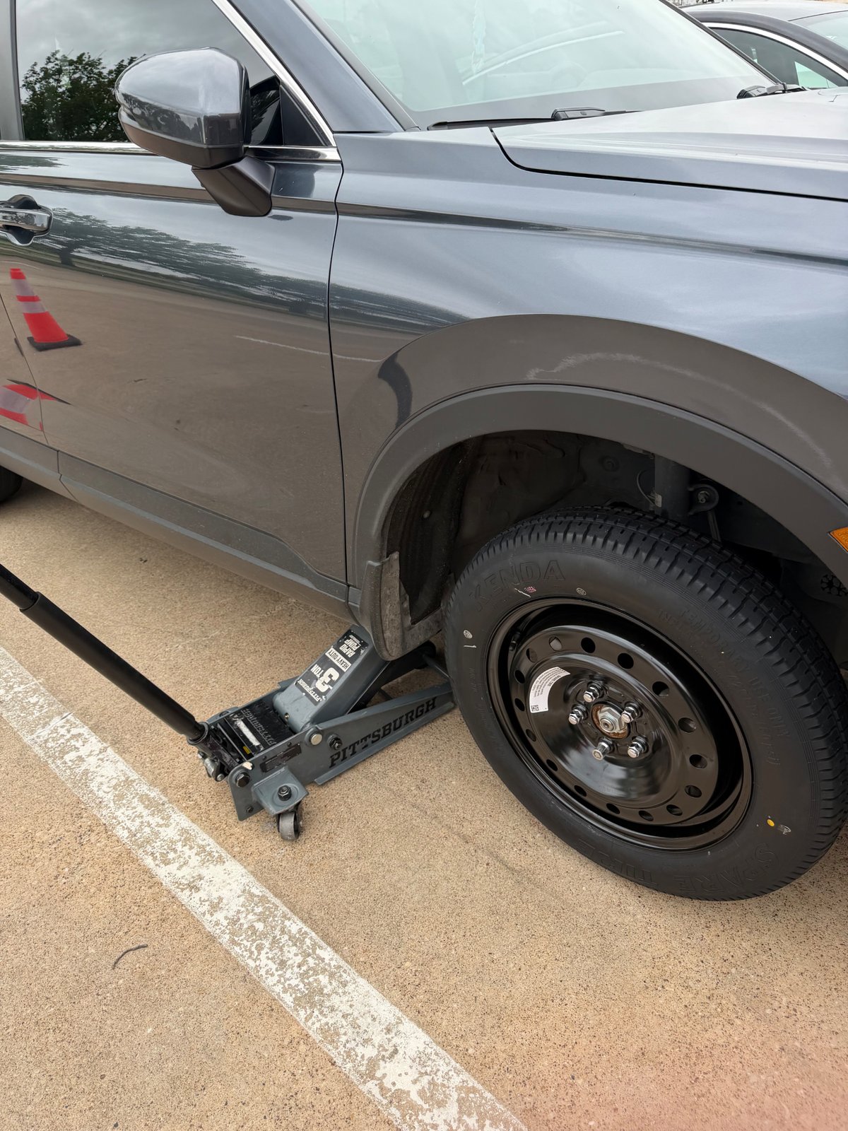 Gray Ford Bronco Sport lifted on a 3-ton Pittsburgh floor jack in a Fort Worth parking lot with a full-size Kenda spare tire mounted, safety cones deployed by All Hours Roadside technician