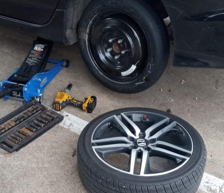 Spare tire mounted on a Honda after a roadside tire swap in Dallas, with floor jack, impact driver, and socket set on the pavement